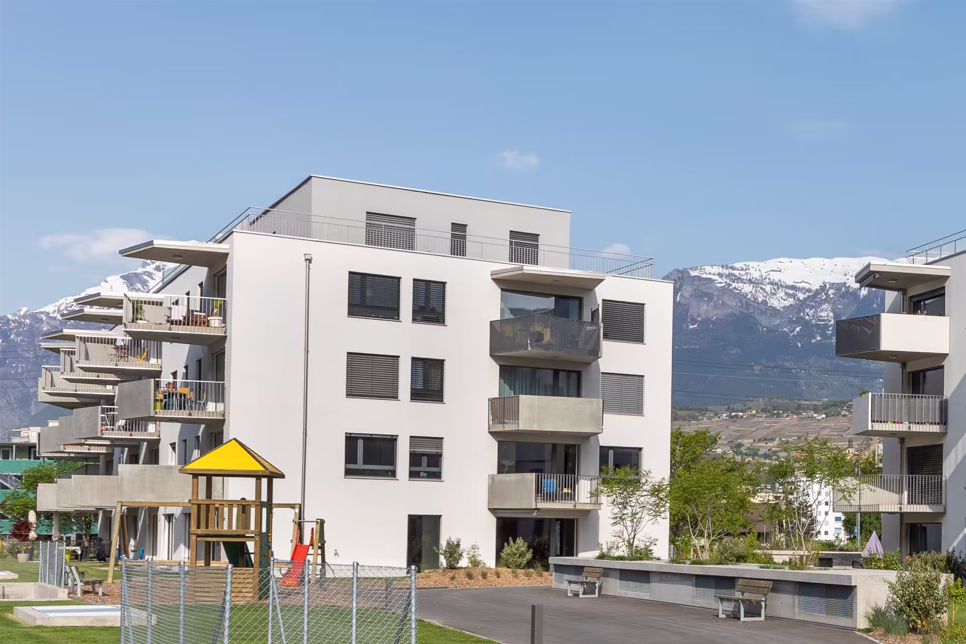 Résidence moderne avec balcons, aire de jeux pour enfants, montagne enneigée en arrière-plan sous un ciel bleu clair.