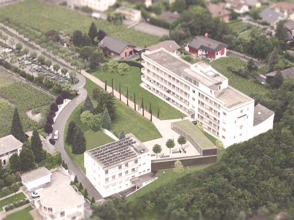 Vue aérienne d’un complexe de bâtiments blancs entourés d’espaces verts, d’arbres, de routes et de plusieurs maisons environnantes.