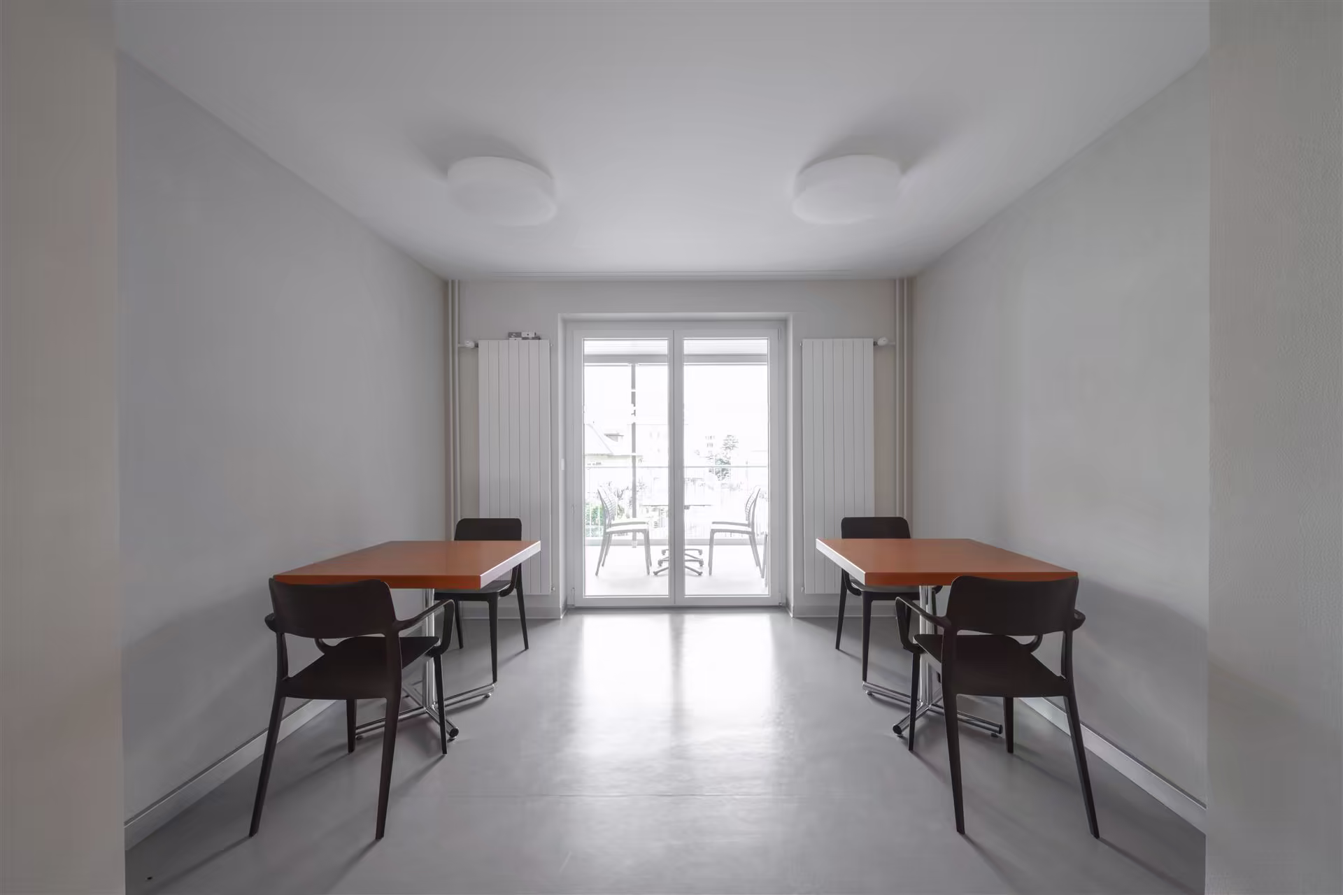 Salle avec deux tables en bois clair, quatre chaises noires, murs gris, sol lisse, portes vitrées donnant sur un balcon avec chaises extérieures.