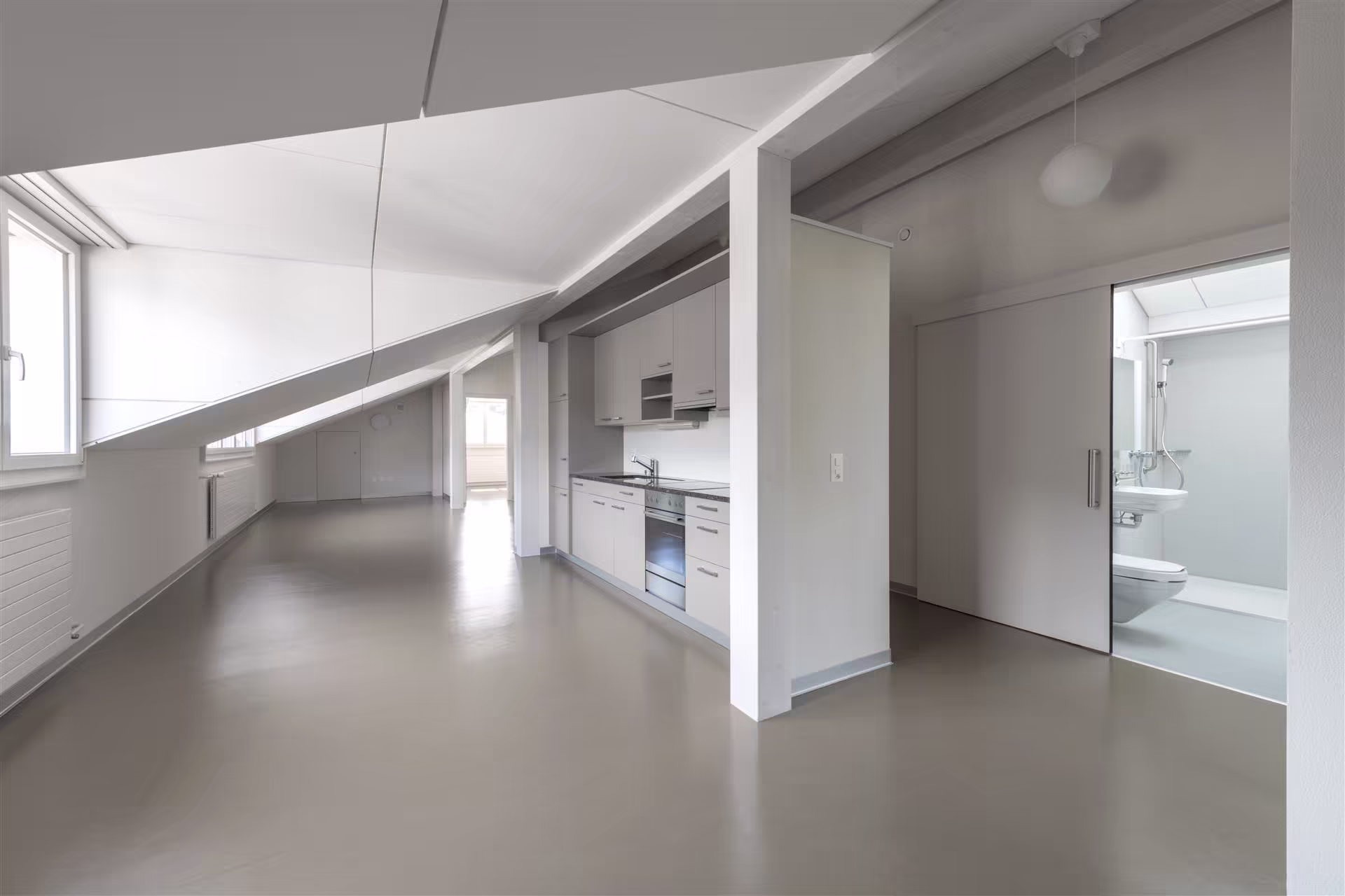 Grand espace intérieur sous pente avec cuisine équipée intégrée, sol gris, murs blancs, fenêtres à gauche et salle de bain visible à droite.