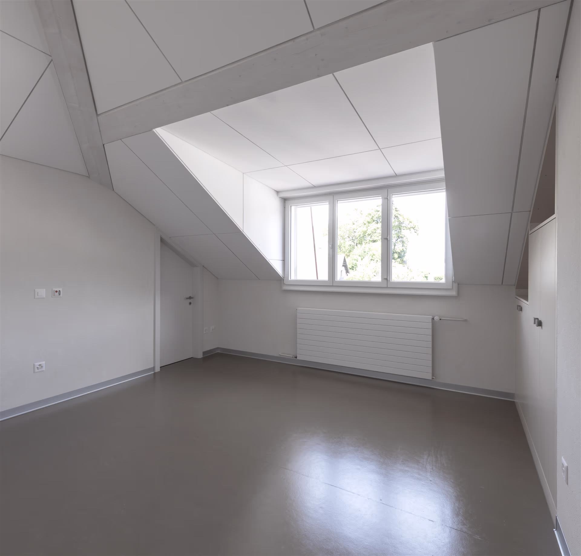 Pièce vide sous les combles avec fenêtre triple, radiateur blanc, sol lisse gris et rangements intégrés sous pente blanche.