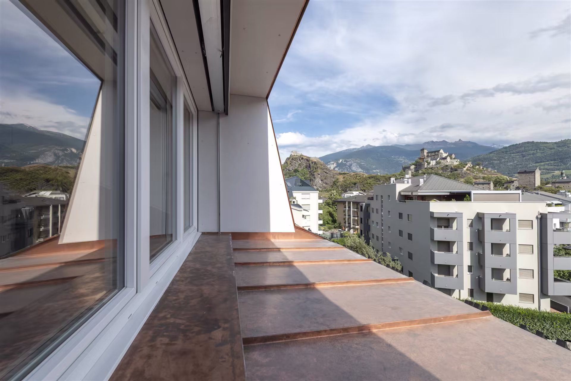 Balcon en métal cuivré avec vue sur des immeubles modernes et un château médiéval sur une colline, montagnes en arrière-plan sous un ciel partiellement nuageux.