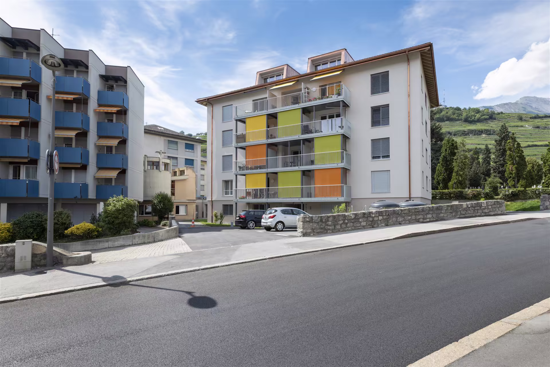 Immeuble résidentiel de quatre étages avec balcons aux panneaux colorés orange, jaune et vert, voitures stationnées, collines verdoyantes en arrière-plan.