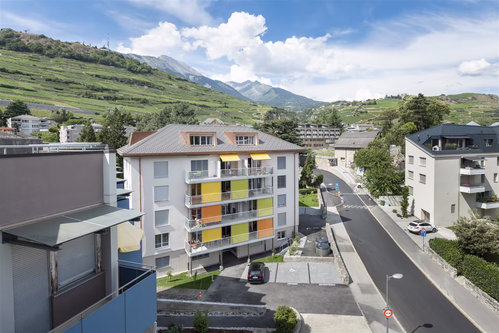 Vue sur un quartier résidentiel avec des immeubles modernes, un parking, une route et des collines verdoyantes en arrière-plan sous ciel bleu éclairé.