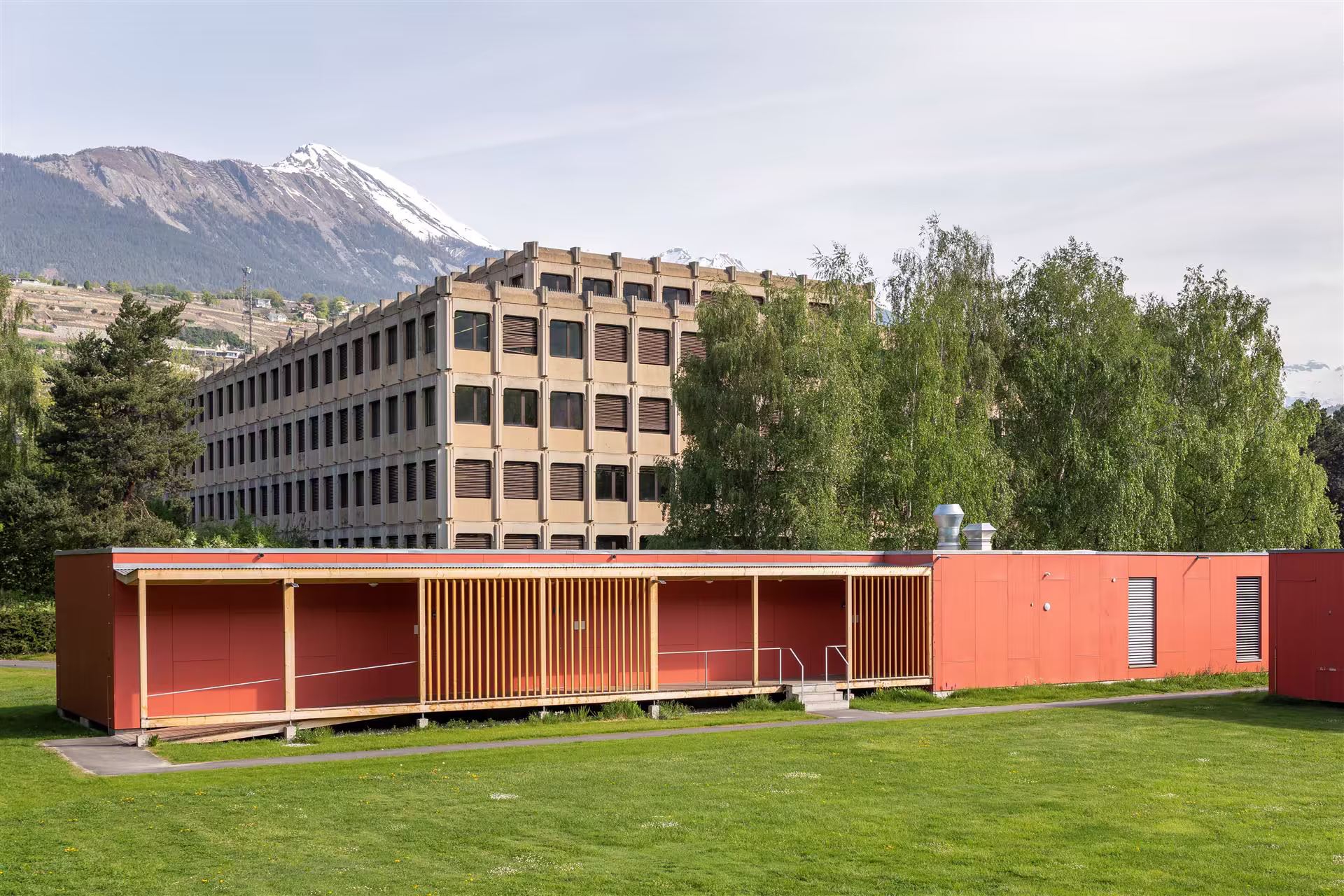 Bâtiment rouge à une étages sur pelouse verte, grands arbres en arrière, immeuble béton à 4 étages et montagnes enneigées au loin sous ciel clair.