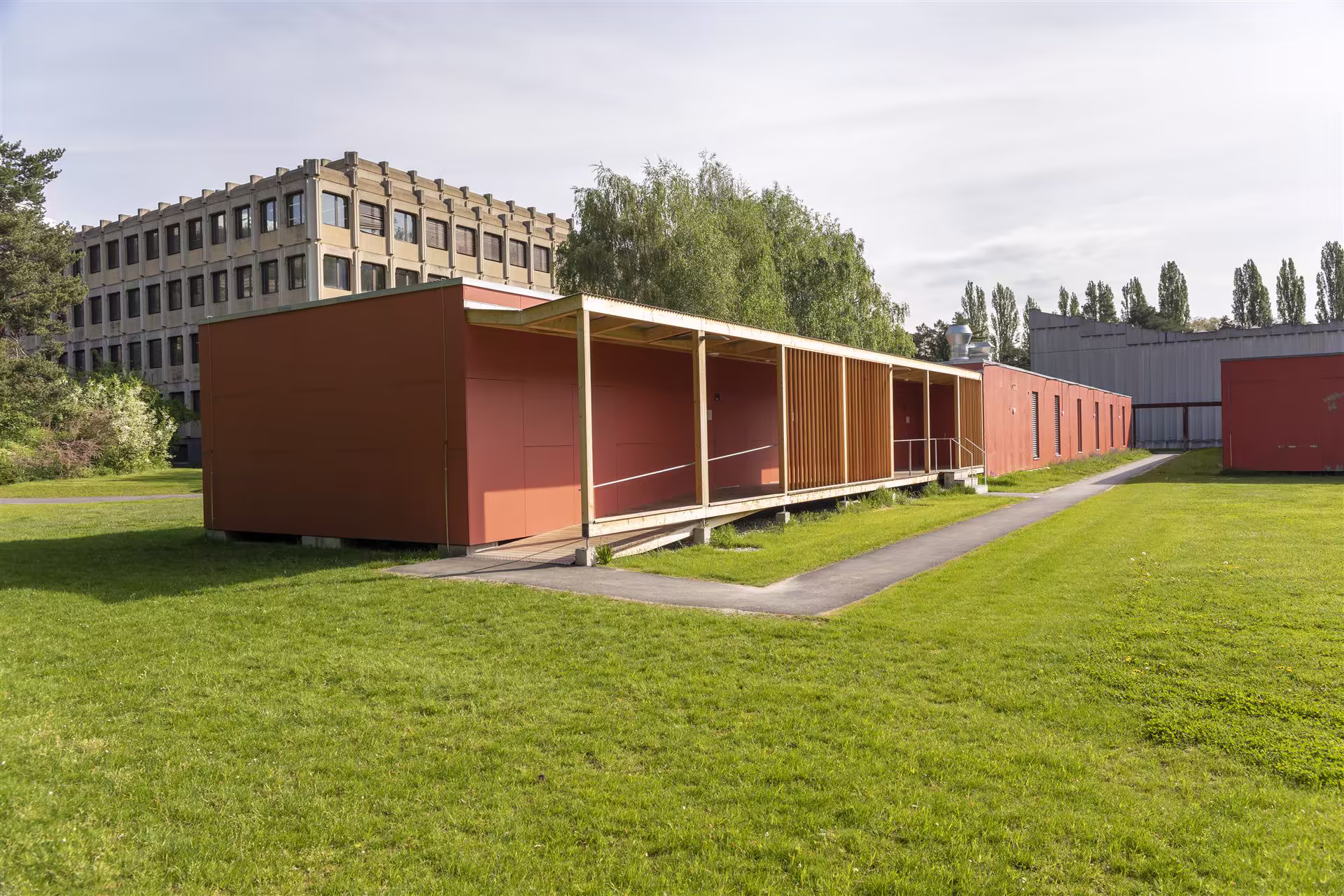 Construction modulaire rouge avec une rampe d'accès, entourée d'herbe verte, arbres, et un grand bâtiment en béton brut en arrière-plan.