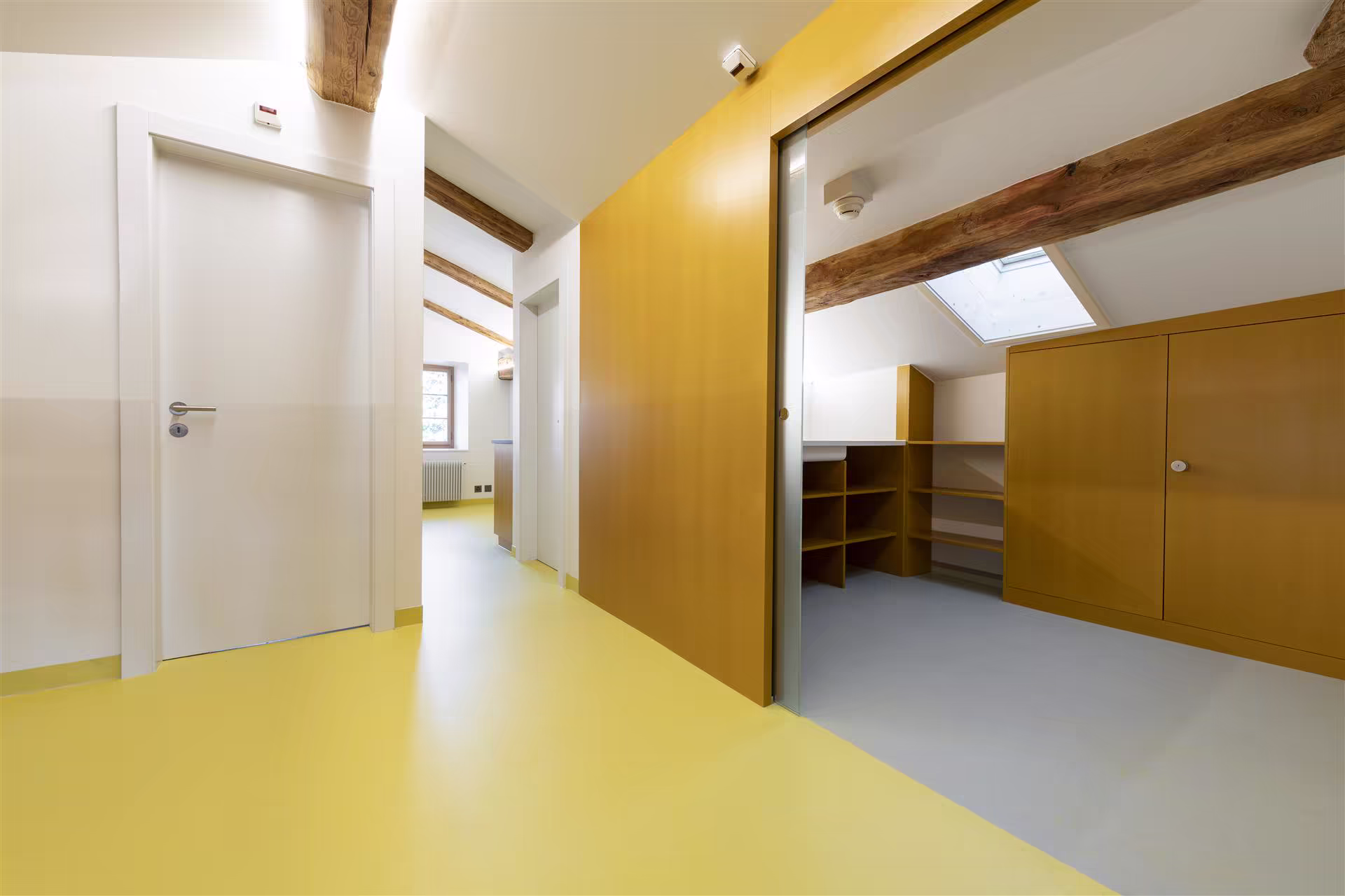 Couloir lumineux avec sol jaune et murs blancs, deux portes blanches à gauche, placard ouvert en bois à droite sous poutres apparentes et velux.