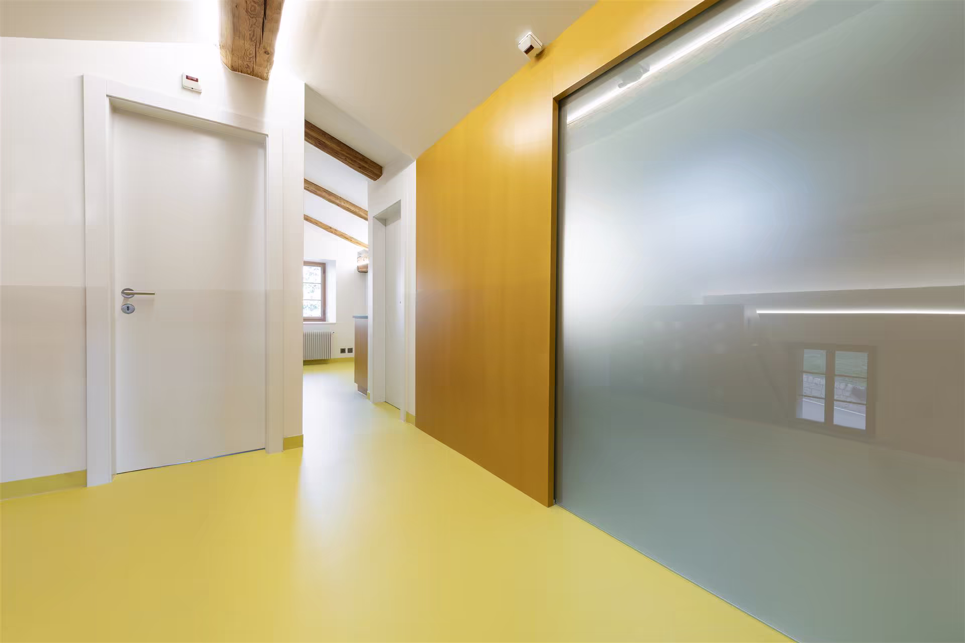 Couloir intérieur avec sol jaune, murs blancs, une porte blanche fermée, une porte coulissante en verre dépoli et poutres apparentes en bois.