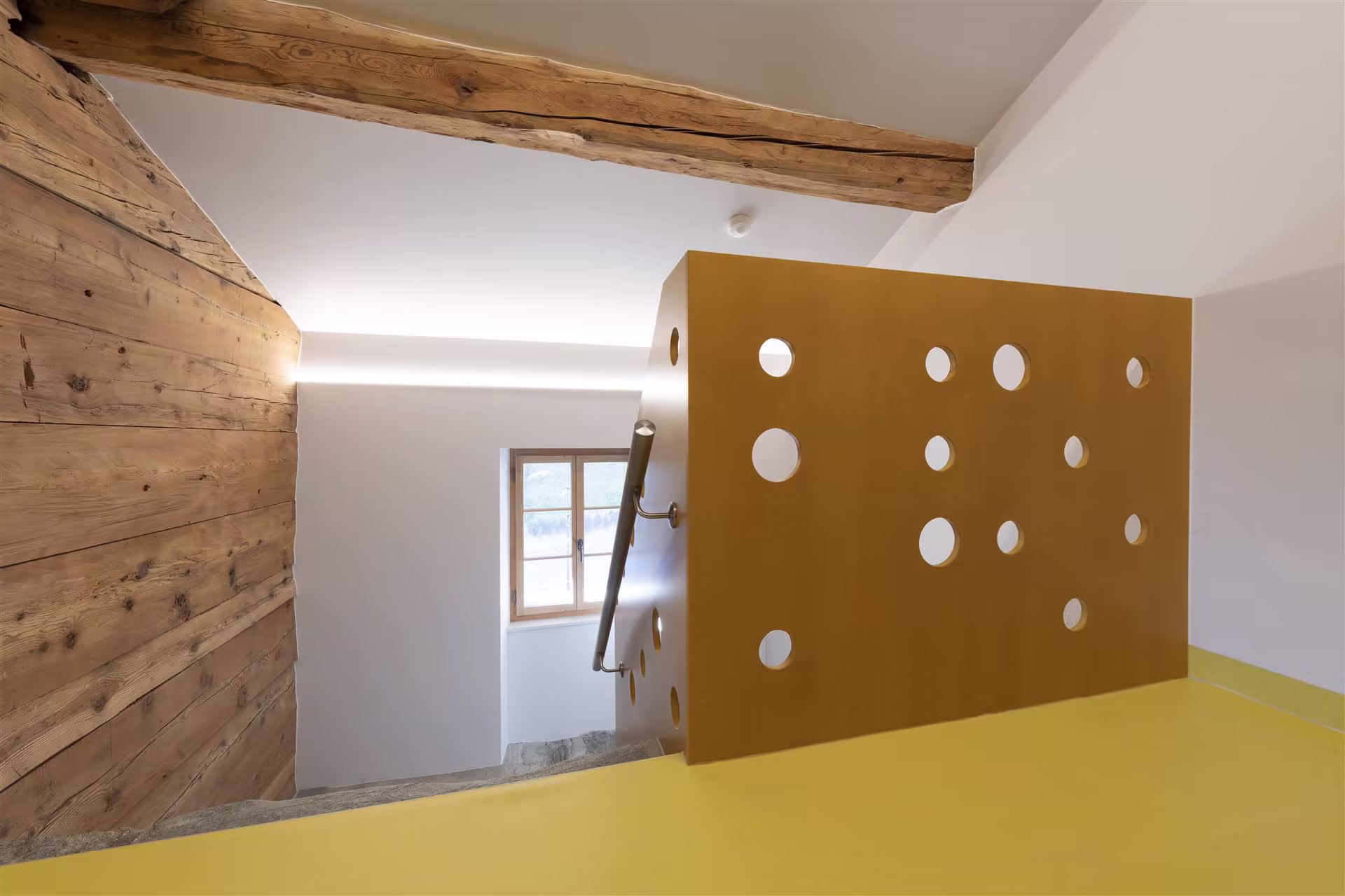 Escalier avec rambarde jaune perforée, murs blancs, mur lambris en bois, poutres apparentes et fenêtre en bois donnant sur l'extérieur montagneux.