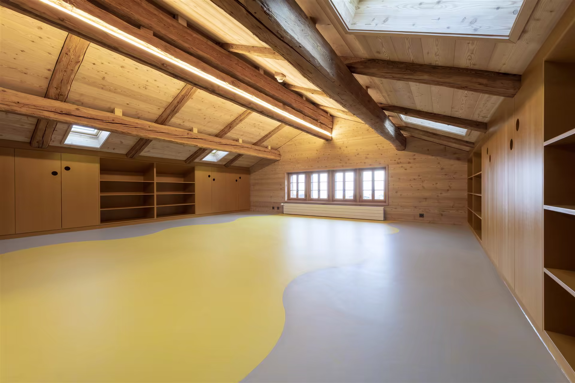 Grand espace mansardé en bois clair, avec poutres apparentes, sol en deux tons jaune et gris, rangements muraux intégrés et fenêtres de toit.