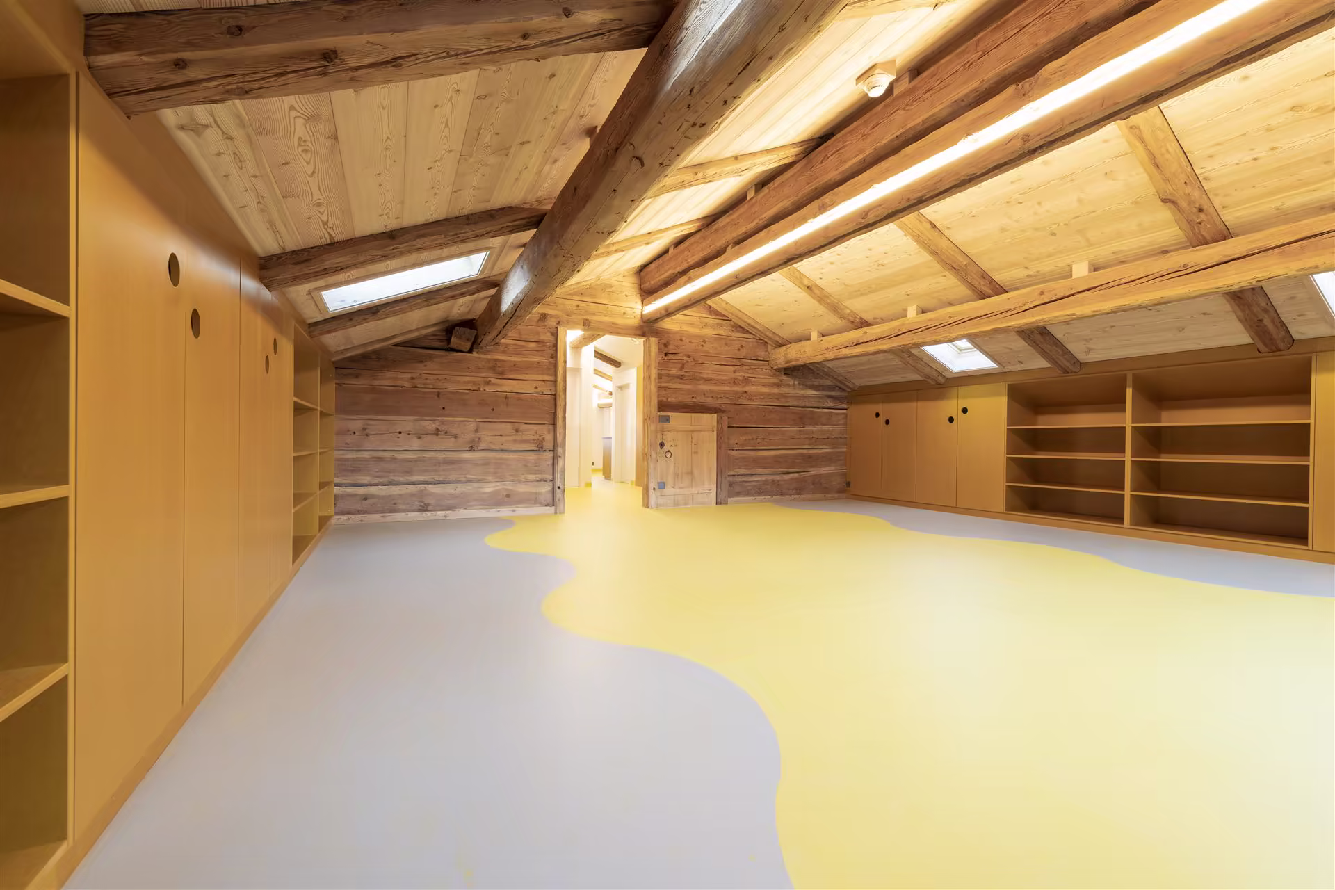 Grande pièce sous combles avec plafond et murs en bois, sol jaune et gris, rangements encastrés le long des murs, deux fenêtres de toit.