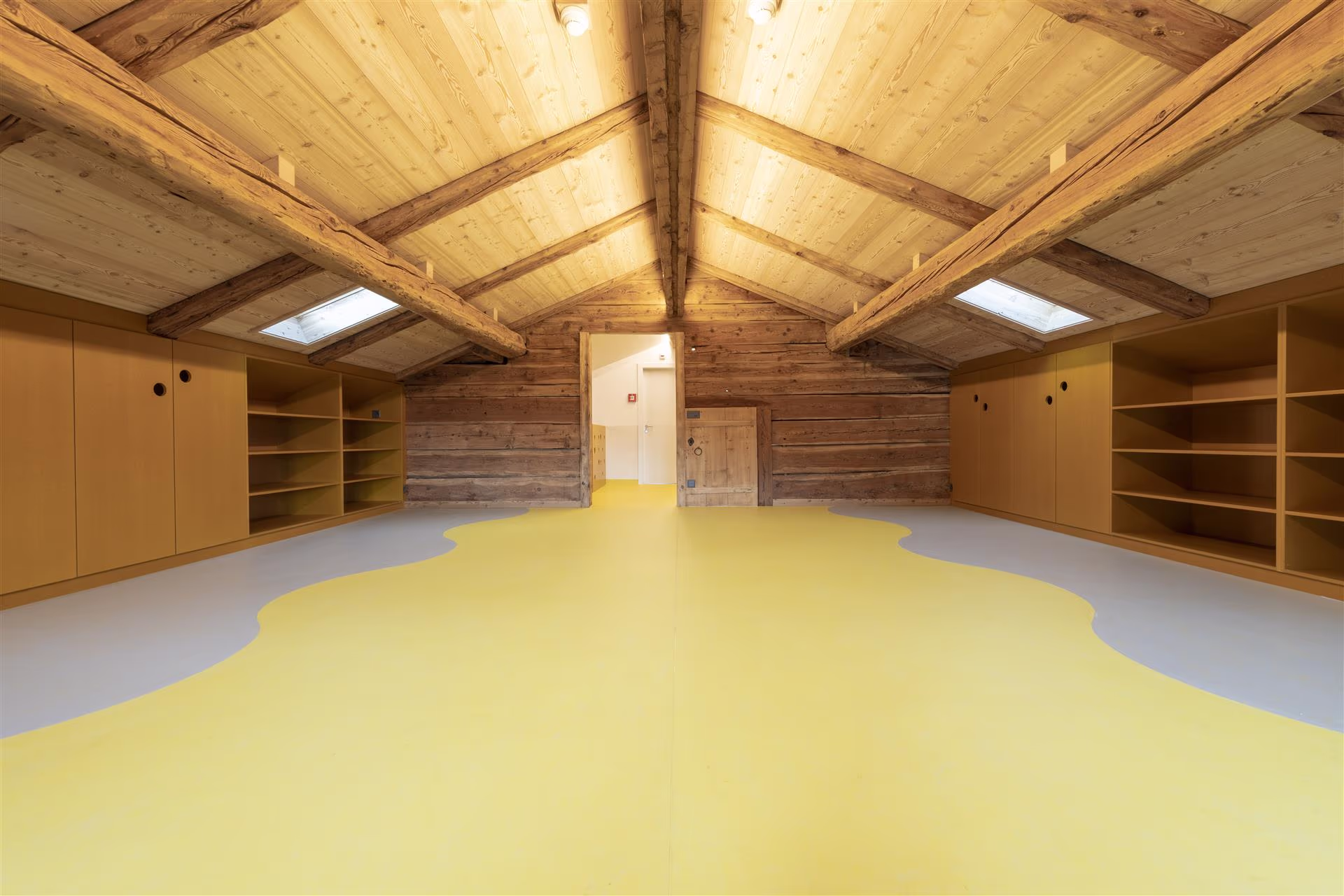 Combles aménagés avec plafond et murs en bois, sol jaune et gris, rangements encastrés, deux fenêtres de toit et porte blanche au fond.