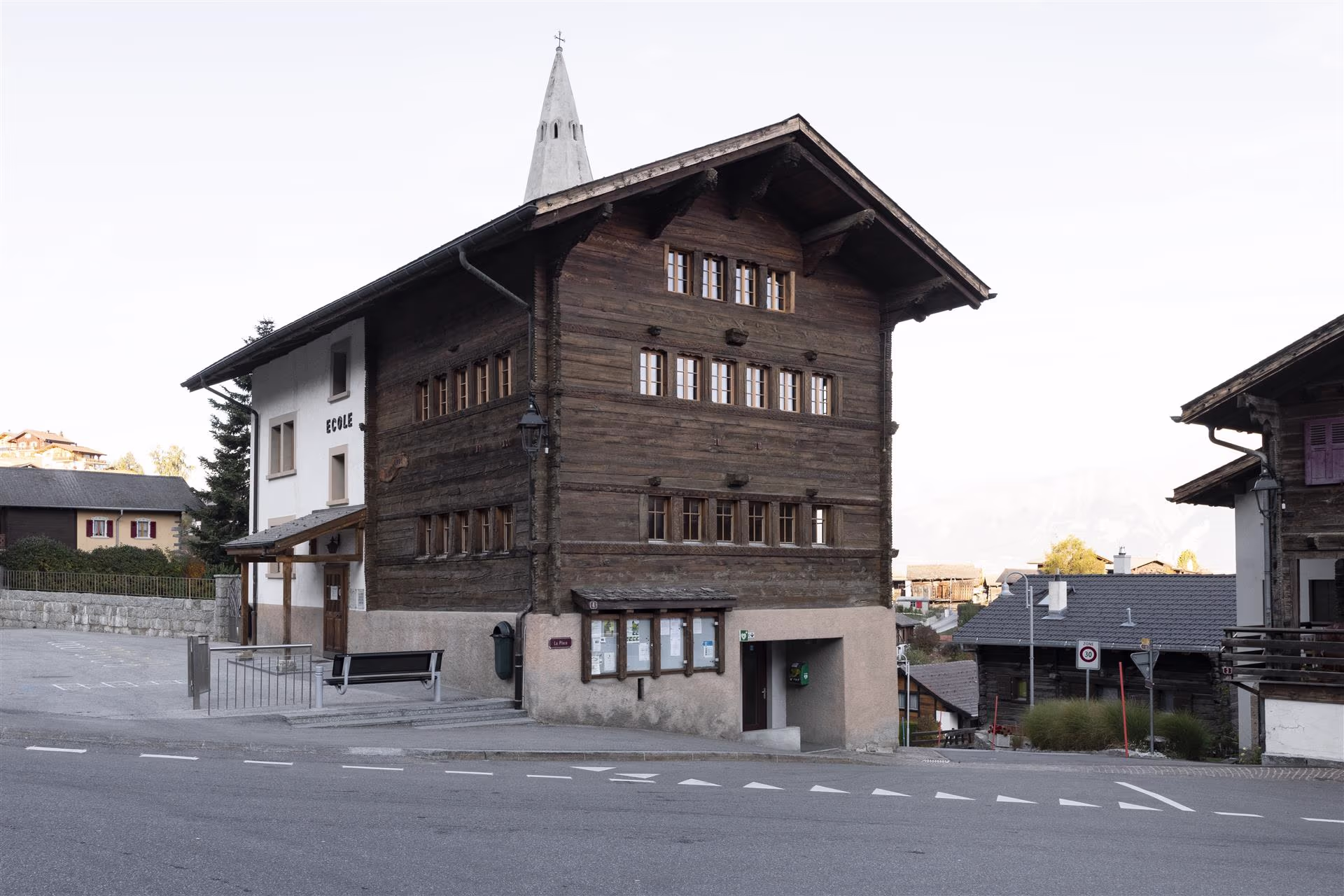 Bâtiment en bois ancien et bâtiment blanc avec l'inscription "ÉCOLE", banc et rues vides dans un village de montagne calme.