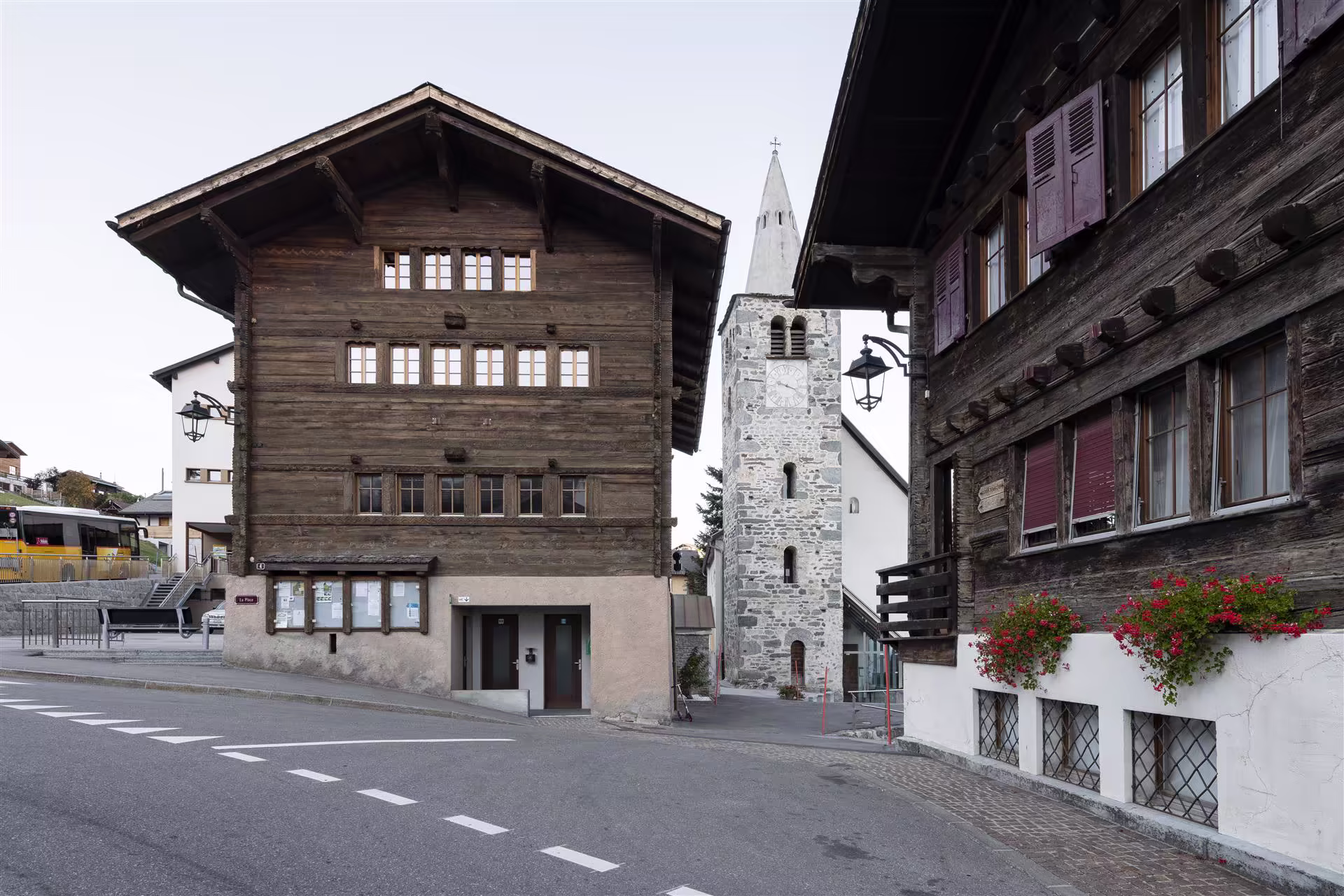 Deux maisons en bois avec fenêtres à volets et balcon fleuri, entre lesquelles se devine un clocher d’église en pierres grises sur une place vide.
