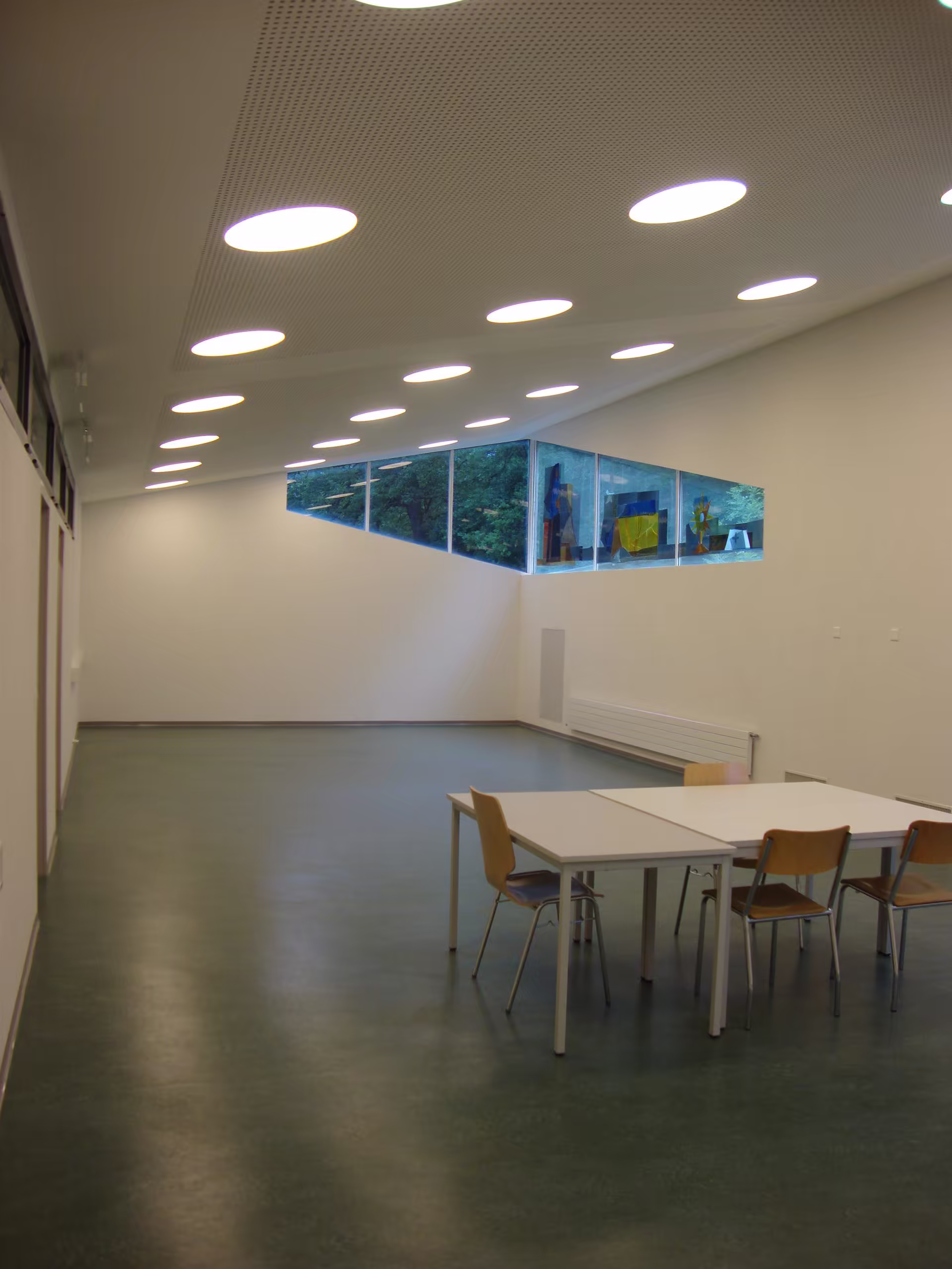 Salle aux murs blancs avec plafond incliné percé de lumières rondes, table blanche et quatre chaises en bois clair, fenêtre triangulaire avec vue sur arbres.