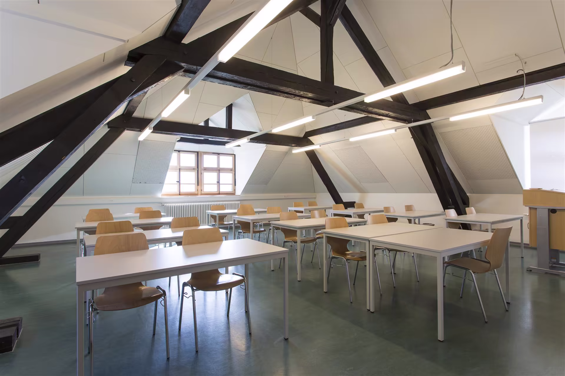 Salle de classe vide sous comble avec poutres en bois noires, tables blanches et chaises en bois clair, éclairage suspendu et fenêtre ouvrant sur l'extérieur.
