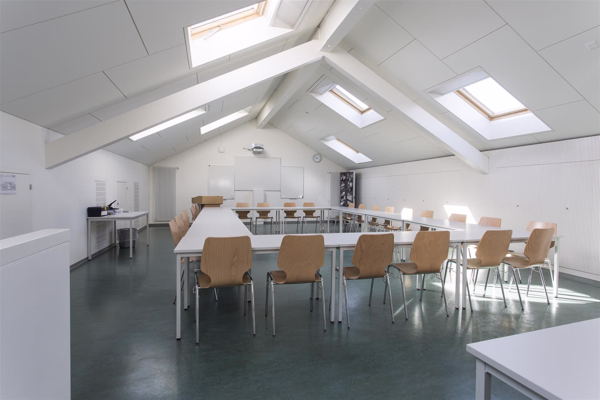 Salle de réunion sous combles avec tables blanches en U, chaises en bois clair, 4 fenêtres de toit et tableaux blancs au mur.