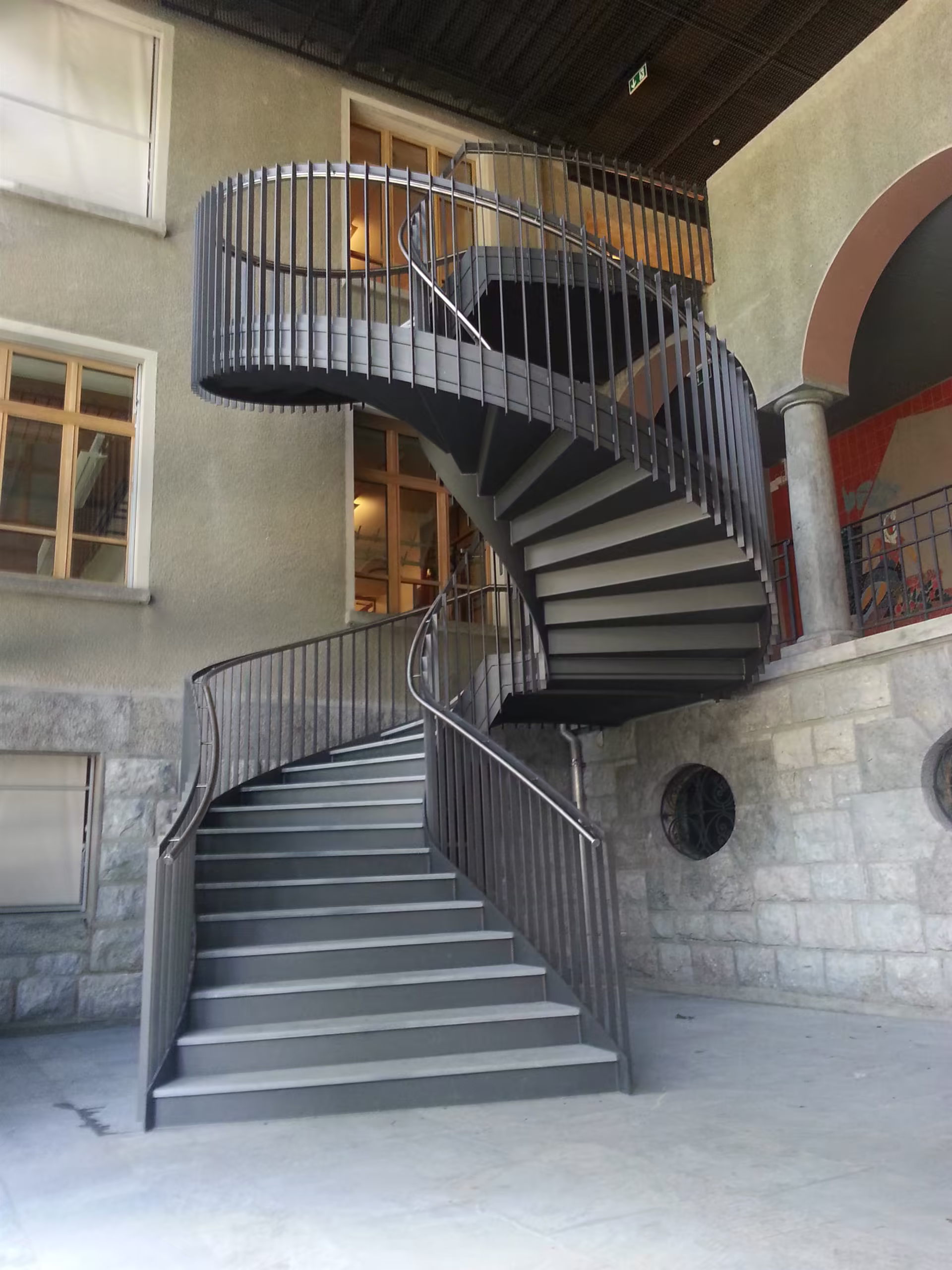 Escalier en colimaçon métallique gris à double balustrade rondes, installé dans une cour intérieure aux murs en pierre et béton.