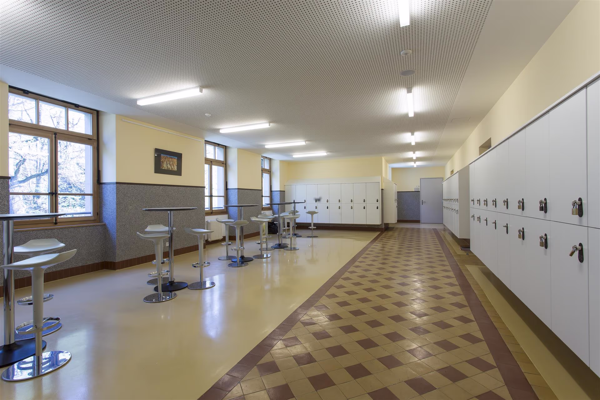 Grande salle lumineuse avec rangées de casiers blancs, plusieurs tables hautes rondes avec tabourets blancs et fenêtres sur un mur.