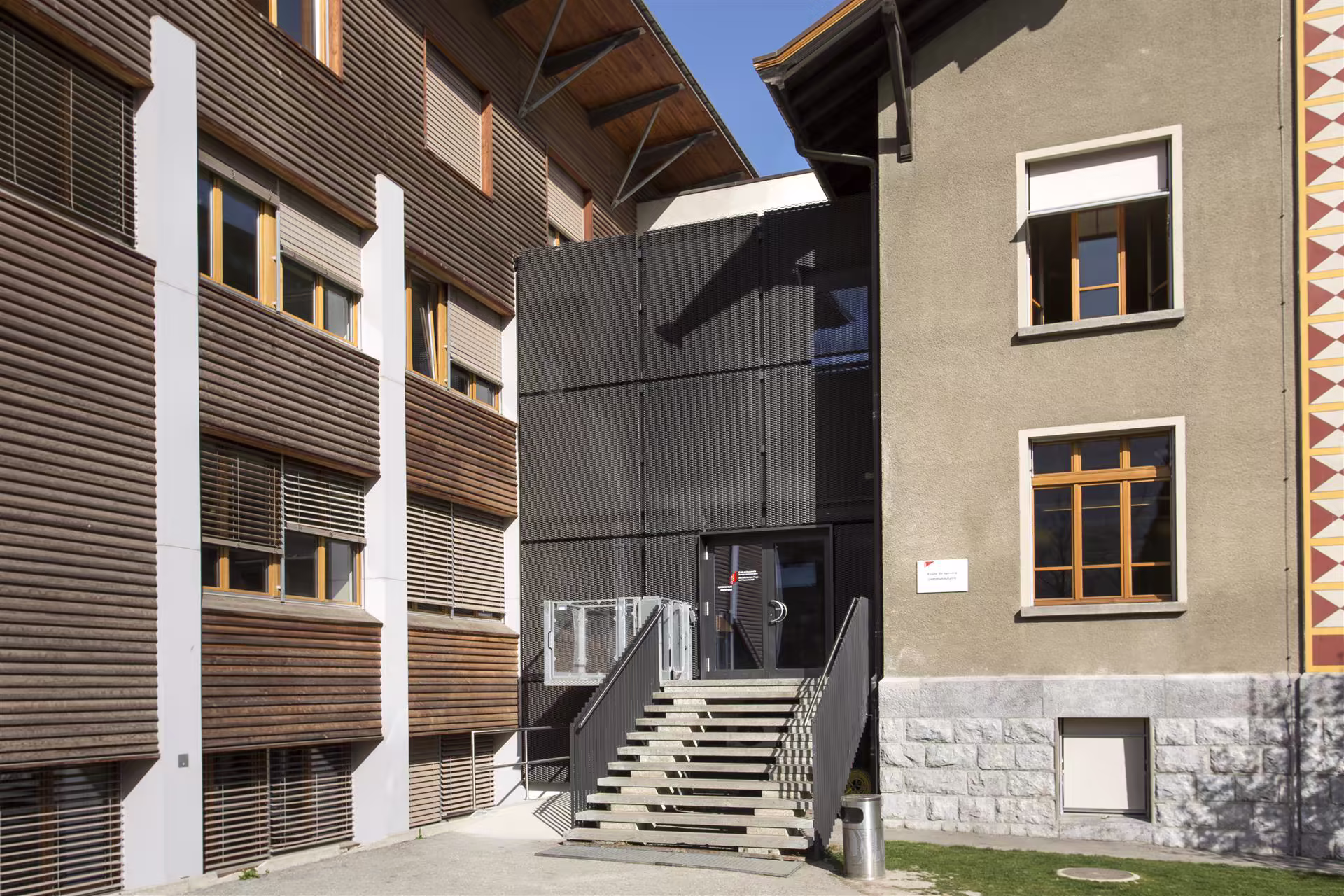 Entrée d’un bâtiment avec escalier extérieur en béton, murs en bois, métal noir et béton, fenêtres en bois avec volets et ciel bleu.