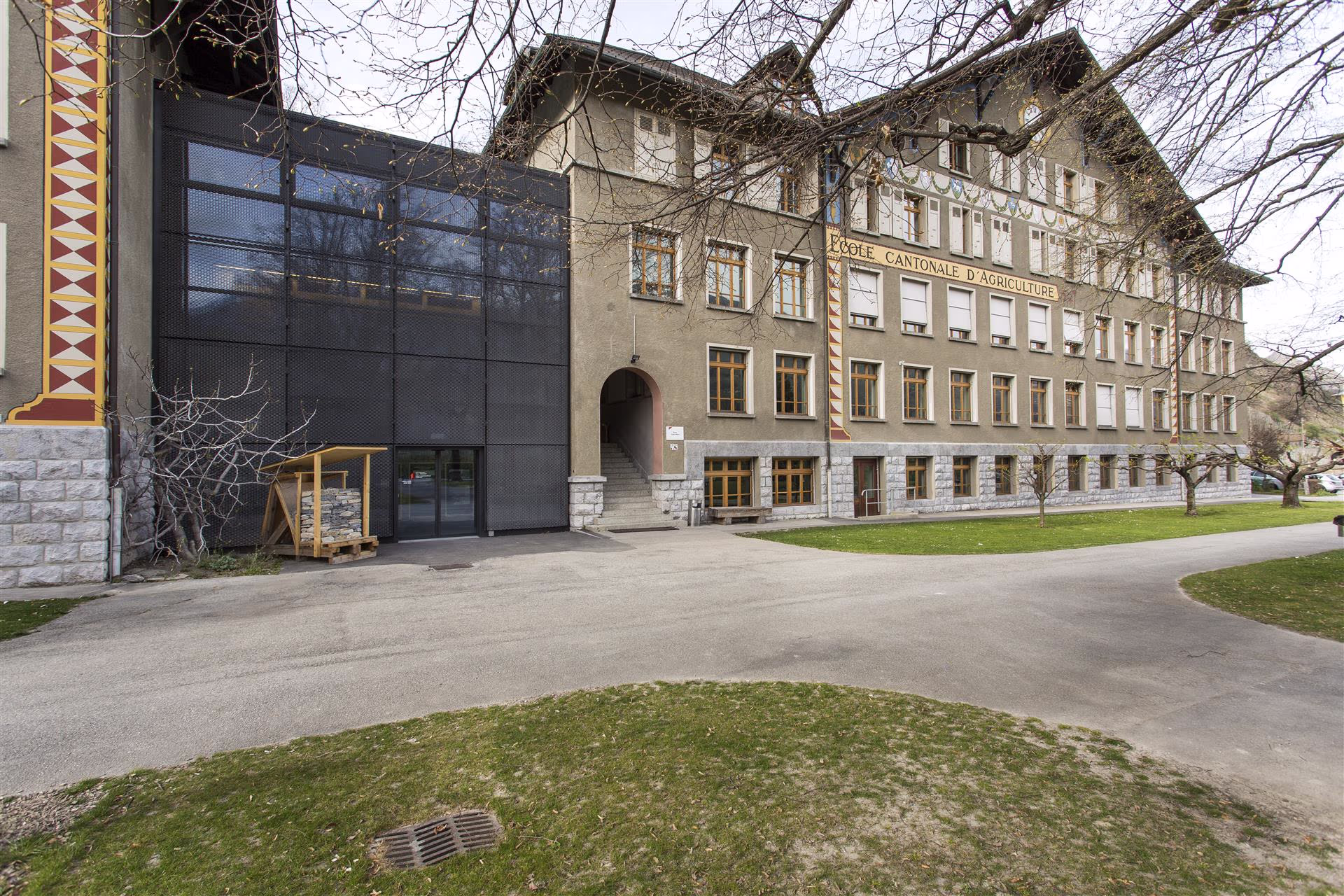 Façade d’un grand bâtiment beige avec inscription "École cantonale d’agriculture", fenêtres en bois, pelouse et arbres sans feuilles au premier plan.