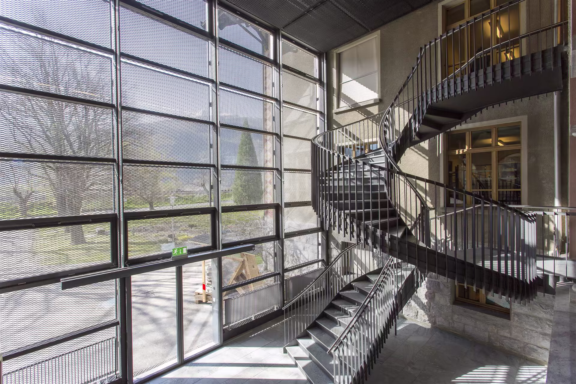 Escalier en métal avec rampe serpentin dans un hall lumineux aux grandes baies vitrées et murs en béton gris. Lumière naturelle filtrée.
