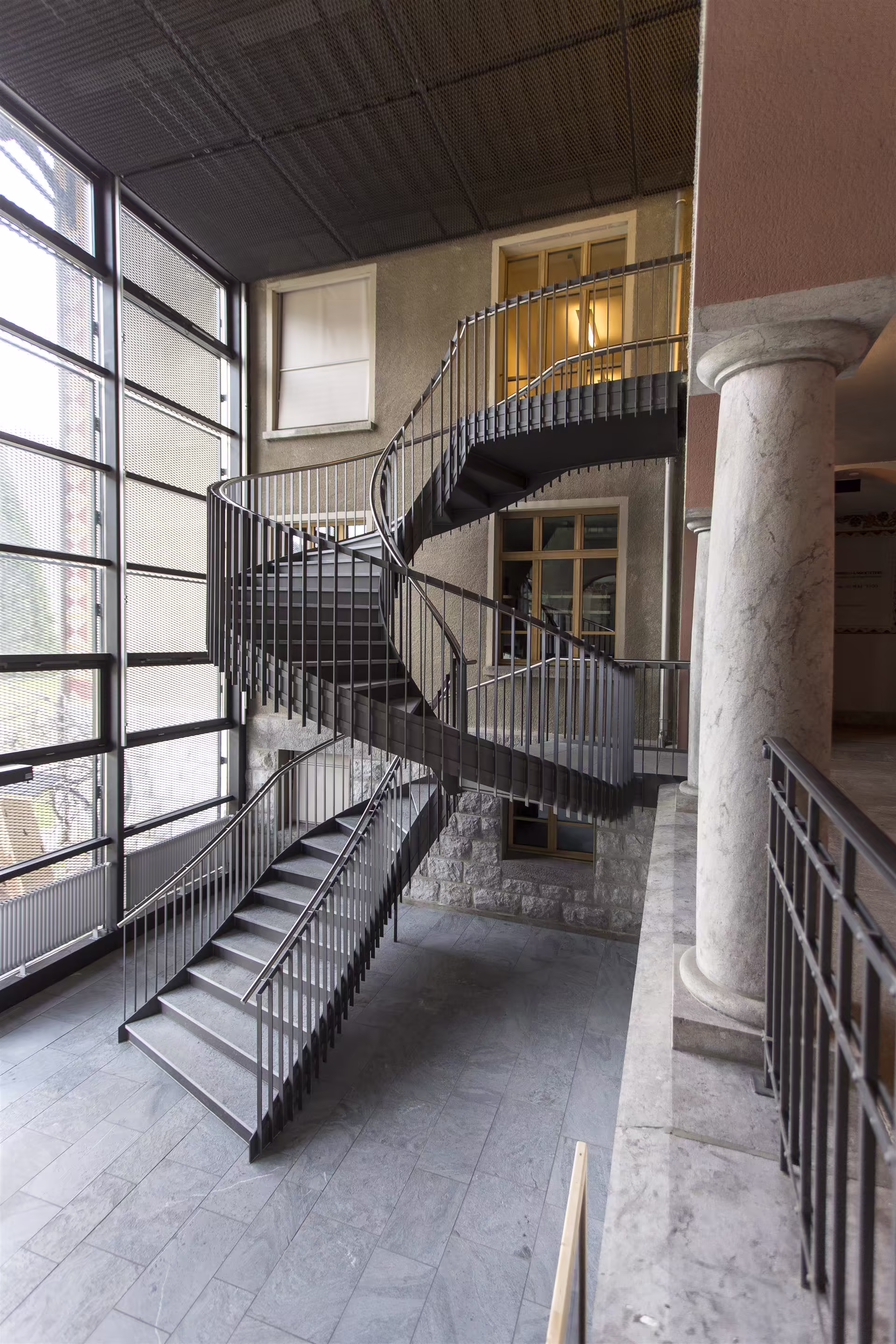 Escalier en métal gris à double hélice dans un espace intérieur avec sol en pierre, grandes fenêtres et colonnes en marbre.