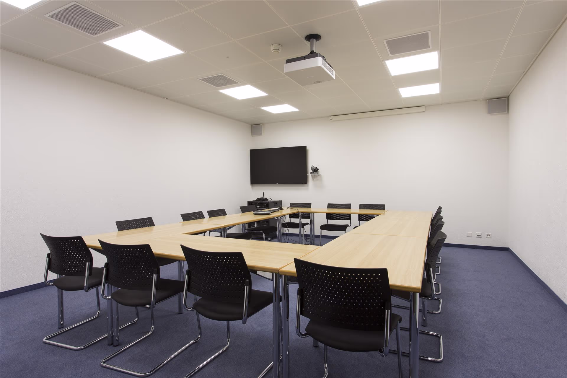 Salle de réunion vide avec tables en U en bois clair, chaises noires, moquette bleue, écran mural et projecteur au plafond.