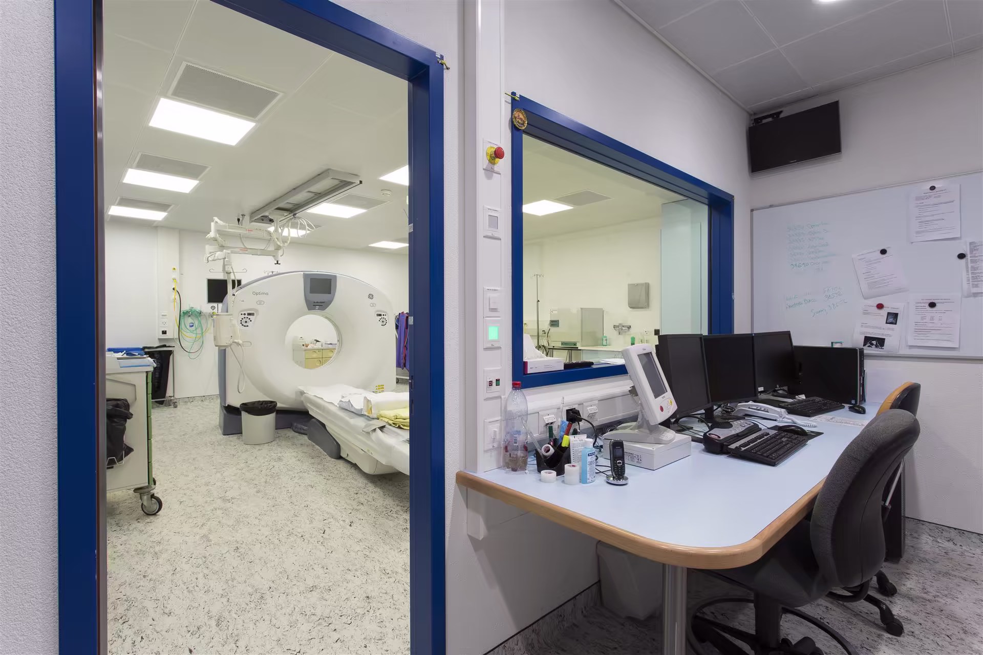 Salle de contrôle d’un scanner médical avec plusieurs écrans et une salle d’examen équipée d’un appareil de tomodensitométrie visible à travers une porte.