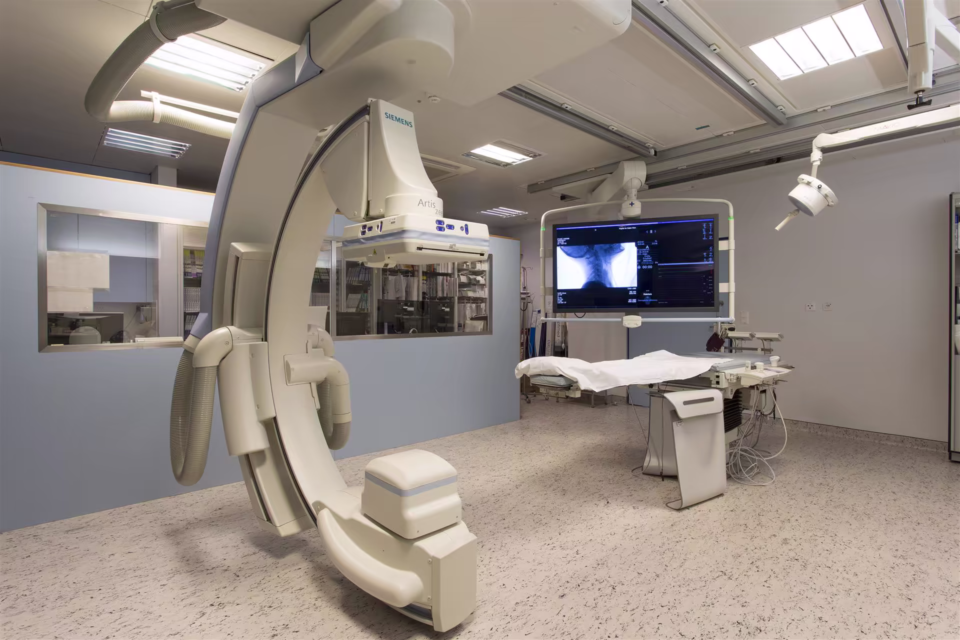 Salle d'examen médical avec un appareil de radiologie Siemens, une table d'examen et un écran affichant une radiographie du cou.