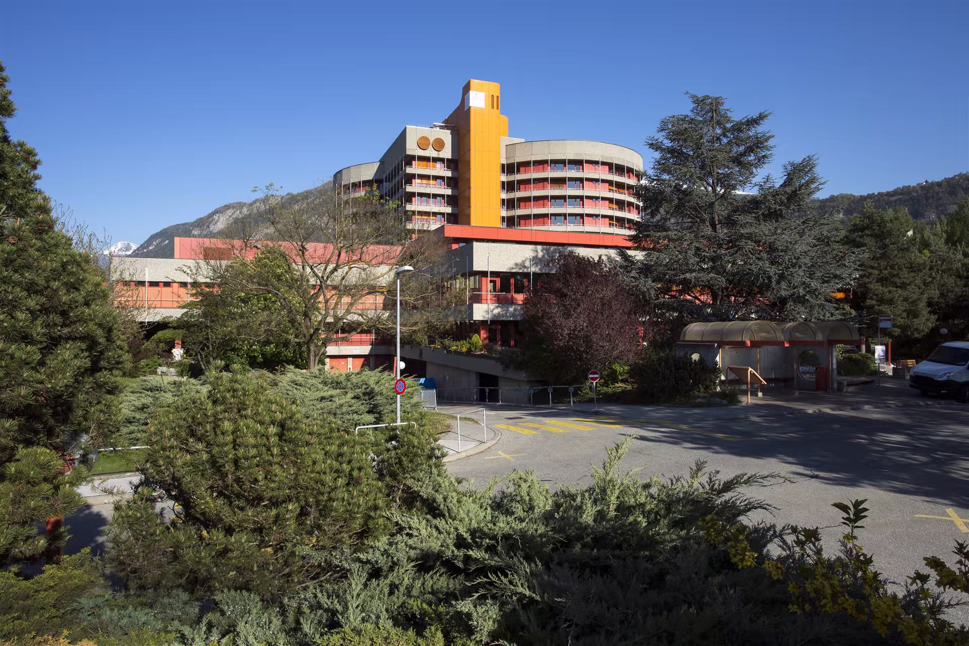 Bâtiment moderne avec une tour orange, entouré d’arbres et jardins, situé dans un cadre montagneux sous un ciel bleu sans nuage.