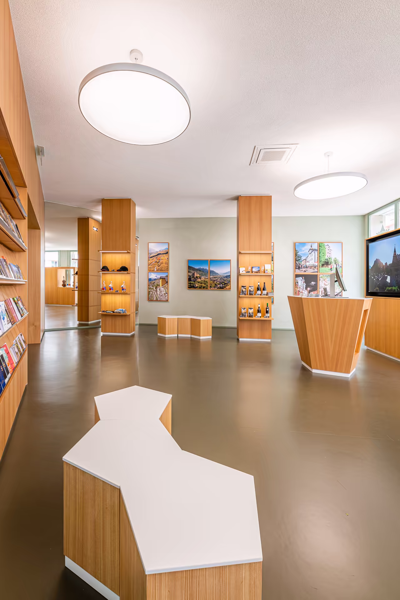 Intérieur lumineux d’un espace d’exposition avec meubles en bois clair, bancs hexagonaux blancs, photographies encadrées aux murs et présentoirs de produits.