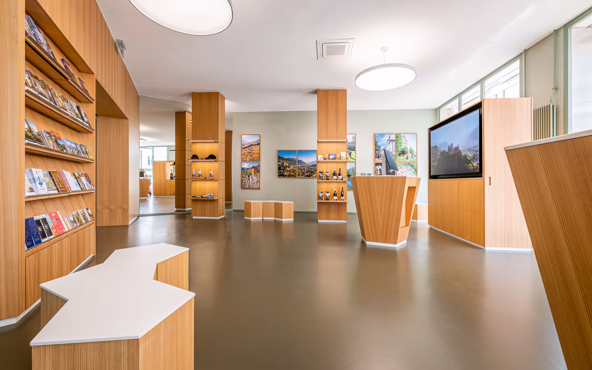 Hall lumineux avec bancs blancs, présentoirs en bois, affiches de paysages aux murs, un comptoir moderne et un grand écran affichant un château en montagne.