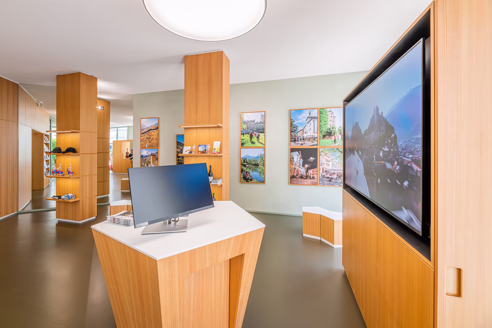 Intérieur lumineux avec mobilier en bois clair, écran d'ordinateur, grand écran mural, et photos encadrées de paysages et villes sur les murs.