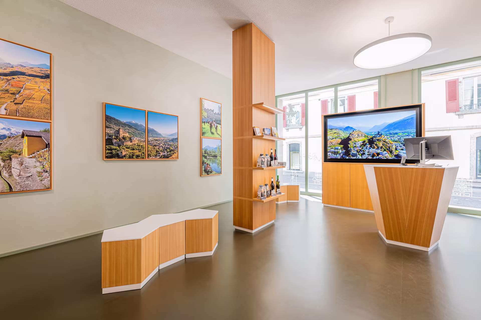 Intérieur moderne avec murs beige, photos de paysages encadrées, mobilier en bois clair, grande TV affichant un paysage de montagnes et vallée ensoleillée.
