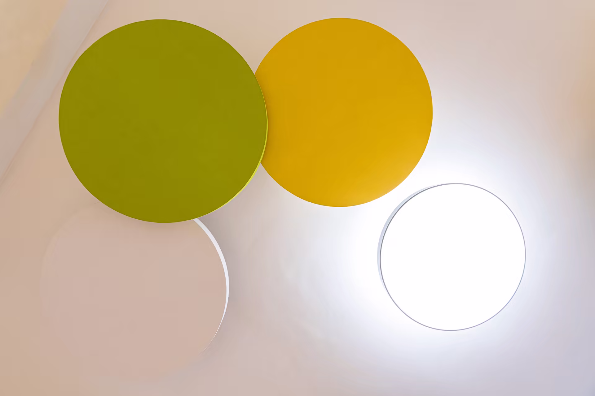 Quatre panneaux ronds suspendus au plafond, en beige, vert, jaune et blanc lumineux, éclairant une pièce aux murs clairs.