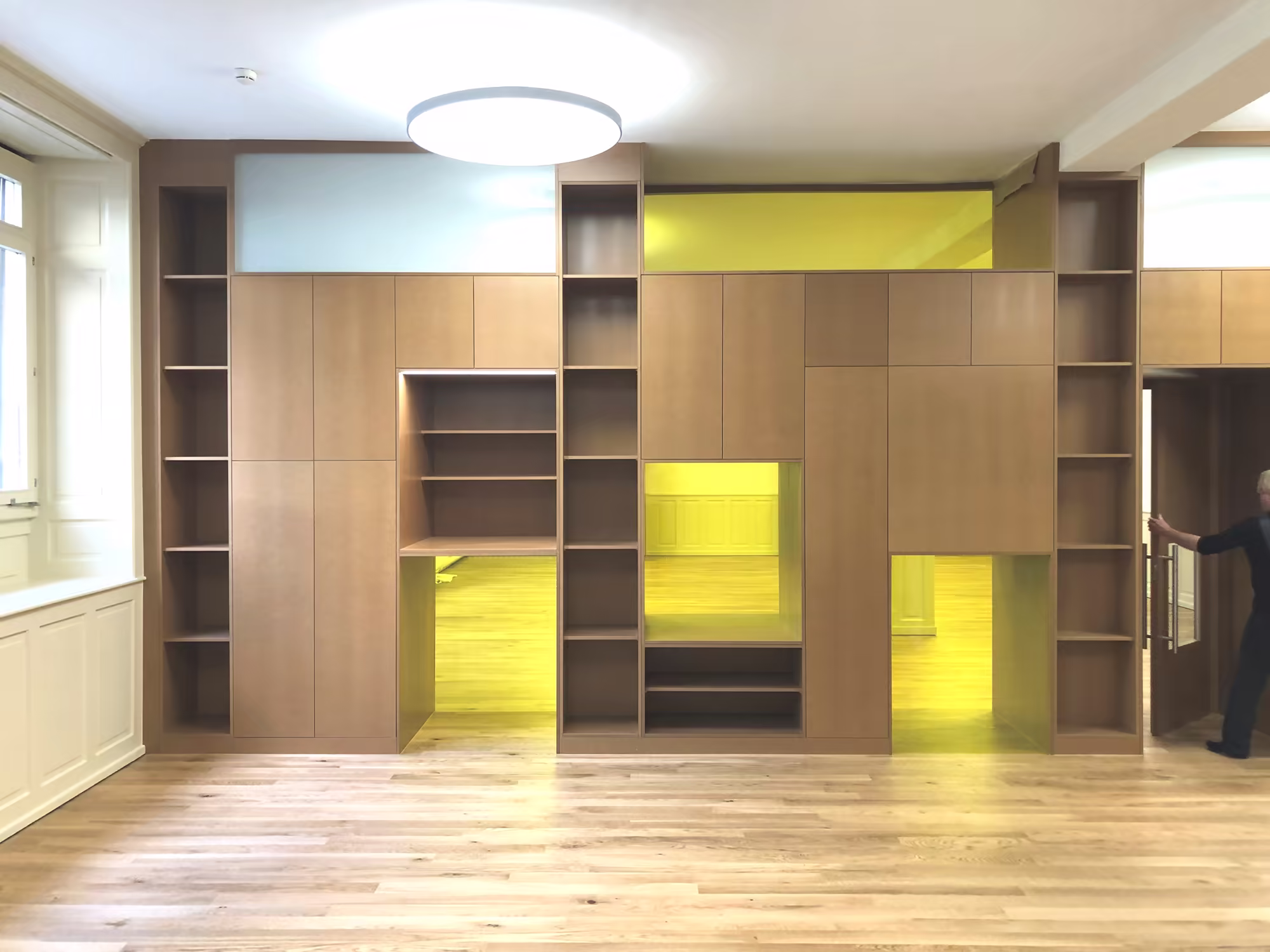 Bibliothèque murale en bois clair avec espaces vides, niches et passages, éclairage jaune à l'arrière, sol en parquet, grande fenêtre à gauche, personne ouvrant une porte à droite.