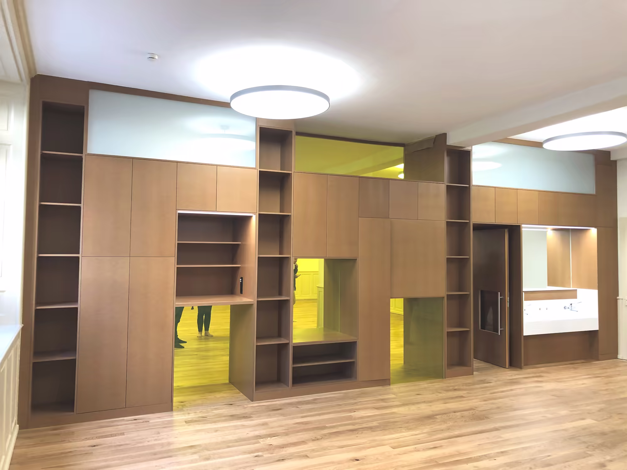 Bibliothèque murale en bois clair avec multiples niches, étagères ouvertes et portes, éclairage circulaire blanc au plafond, sol en parquet clair.