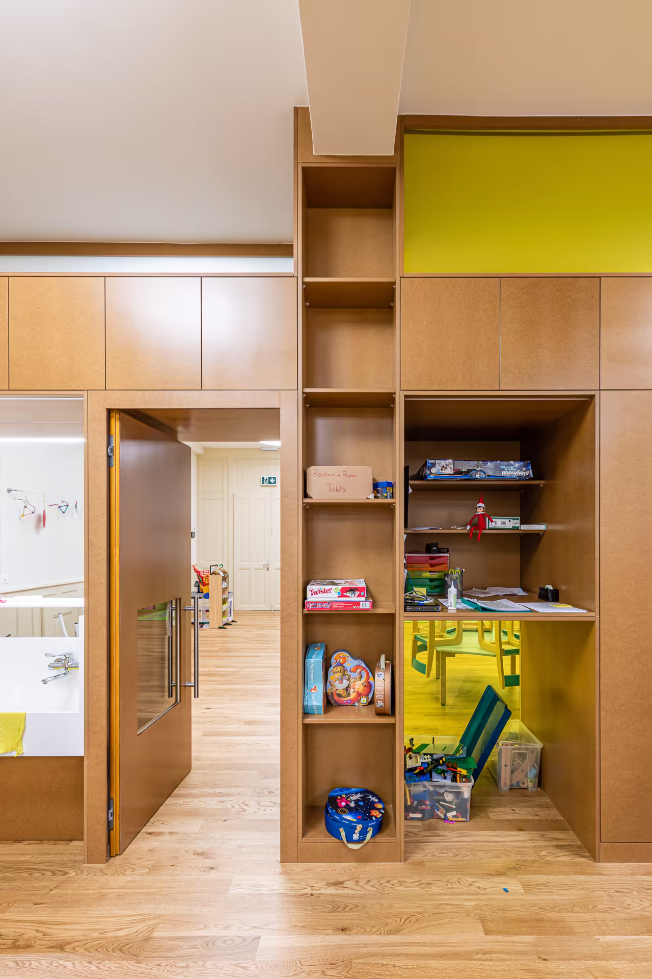 Mobilier en bois clair avec étagères, porte ouverte vers couloir, accès à un espace de jeu avec chaises et jeux de société.
