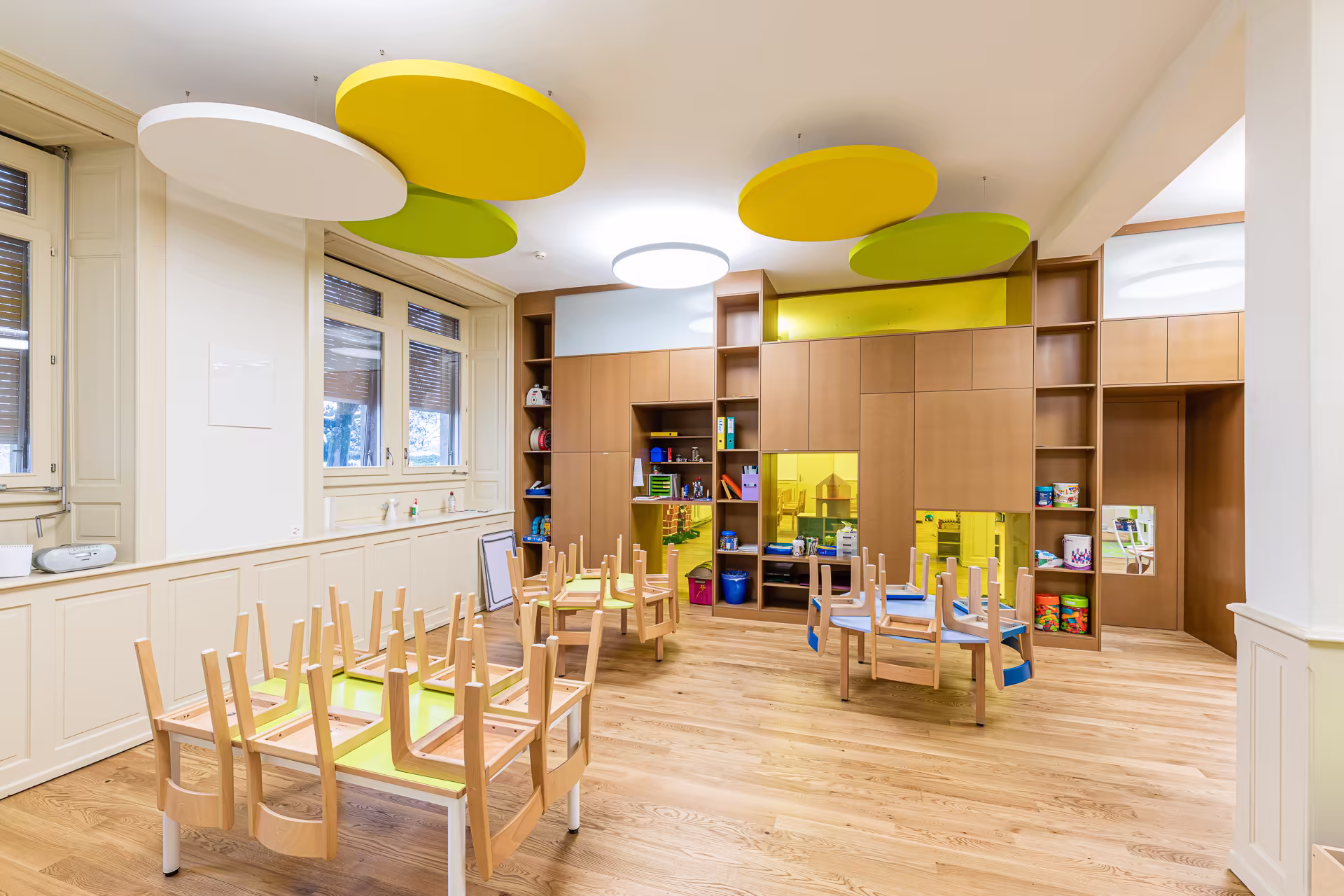 Salle de classe colorée avec tables et chaises en bois retournées, rangements muraux, fenêtres, et panneaux acoustiques jaunes et verts au plafond.