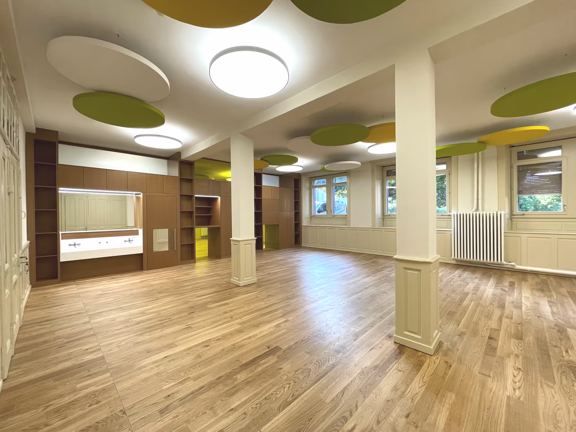 Grande salle lumineuse avec parquet clair, murs beige, colonnes, fenêtres, radiateur, luminaires ronds au plafond et panneaux acoustiques colorés.