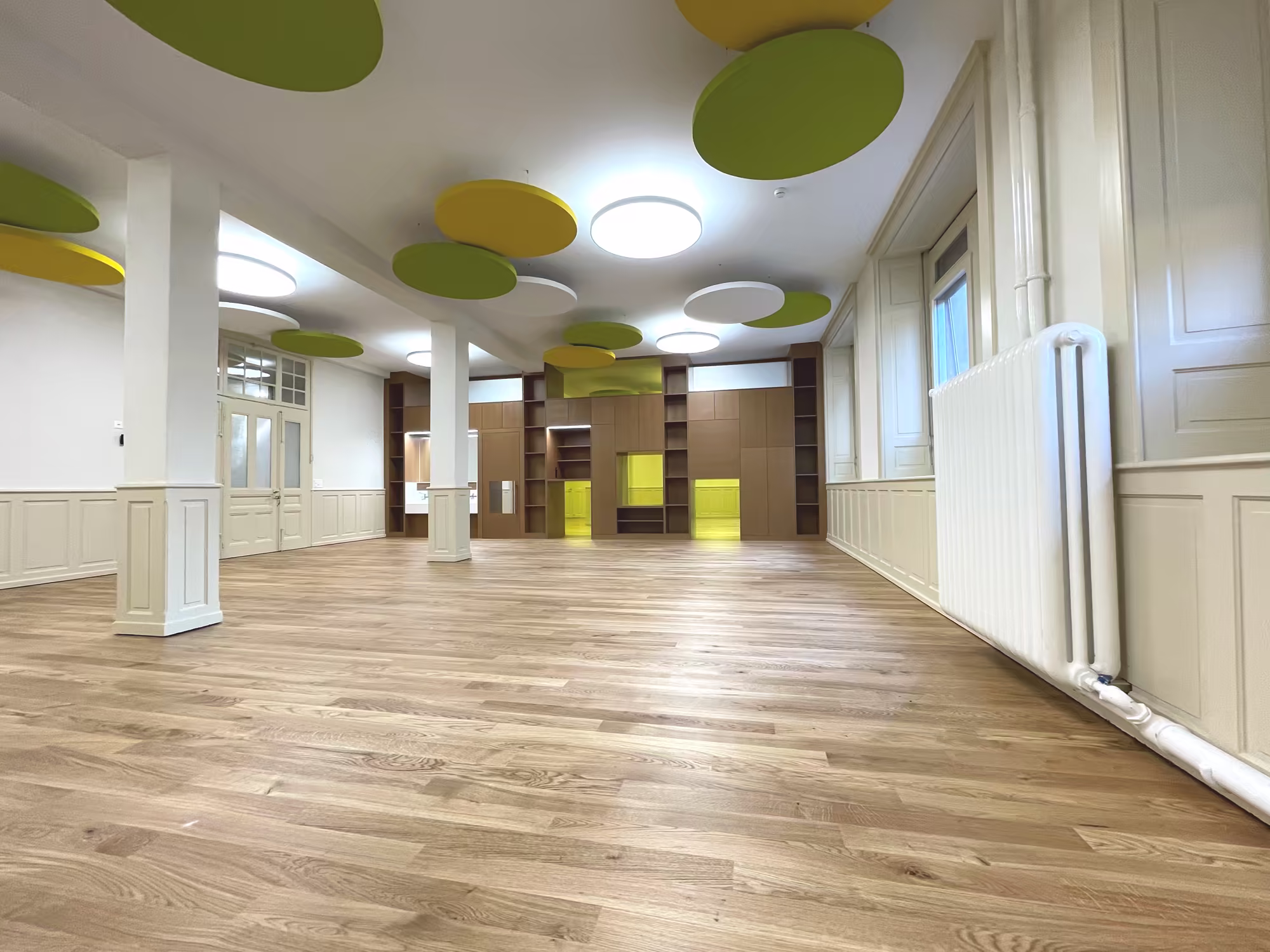 Grand espace intérieur vide avec sol en parquet clair, murs blancs, colonnes, grandes fenêtres, radiateur et décorations circulaires vertes et jaunes au plafond.