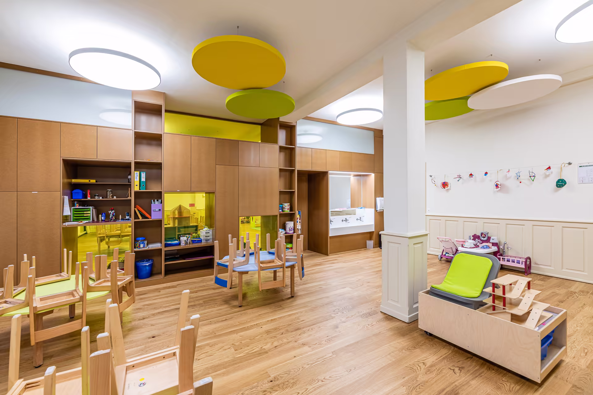 Salle de classe maternelle avec tables et chaises retournées, étagères en bois, jeux, lavabos, et suspensions colorées au plafond.
