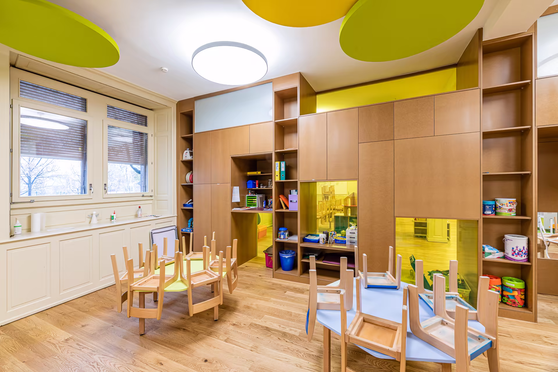 Salle de classe lumineuse avec tables et chaises en bois retournées, grandes fenêtres, rangements muraux marron et décoration jaune et verte au plafond.