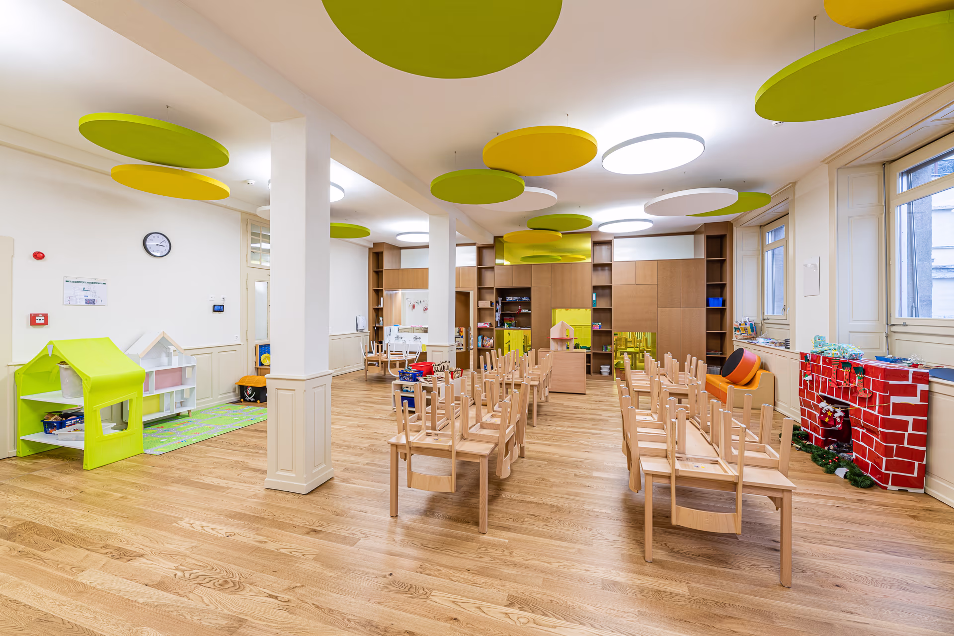 Salle de classe pour enfants avec chaises retournées sur tables, jouets, tapis coloré, meubles en bois clair et décorations circulaires vertes et jaunes au plafond.
