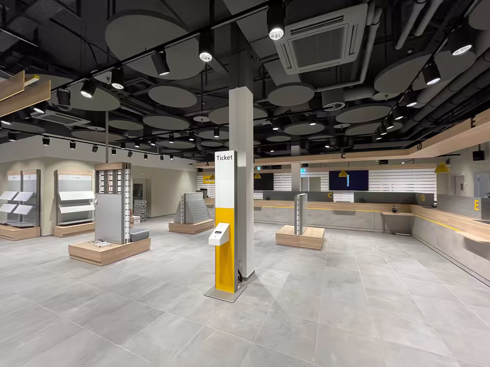 Grand espace intérieur d’un bureau de poste moderne, avec bornes à tickets, comptoirs numérotés et présentoirs vides, éclairage LED et sol en carrelage gris.