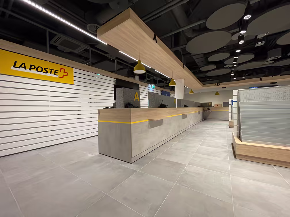 Grand comptoir d’accueil moderne avec lumière jaune, panneaux suspendus en bois clair, sol carrelé gris, et logo La Poste sur un mur blanc à lattes horizontales.