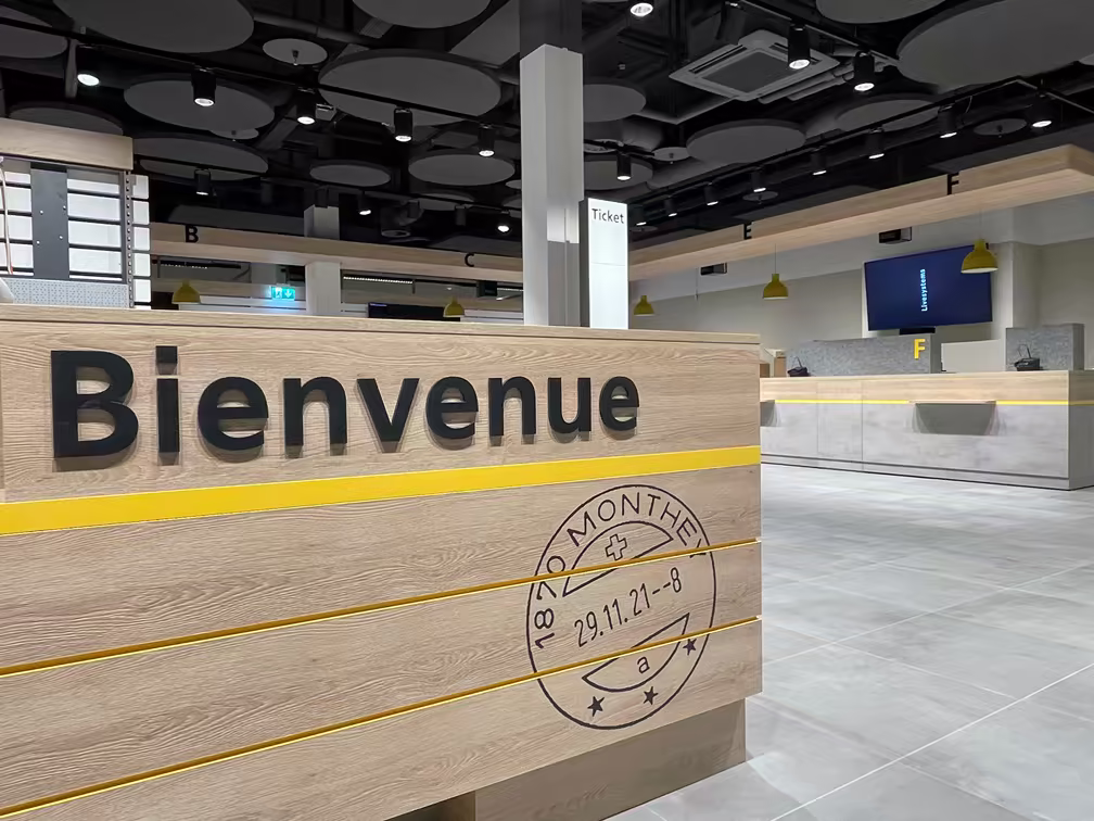 Accueil avec panneau "Bienvenue" en bois clair et lettres noires, comptoirs numérotés en gris, lampe jaune suspendue dans un espace moderne et épuré.