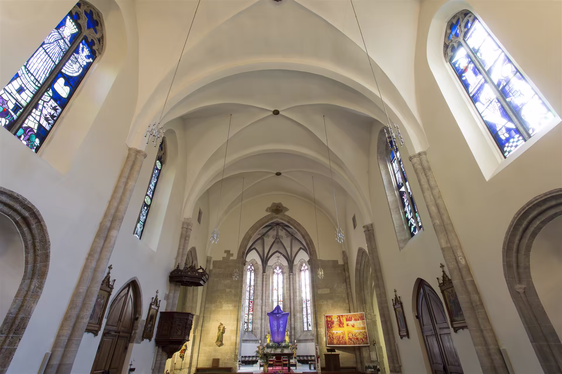 Intérieur d'une église avec voûtes en ogive, vitraux colorés, statues religieuses et autel central décoré de tissu violet.