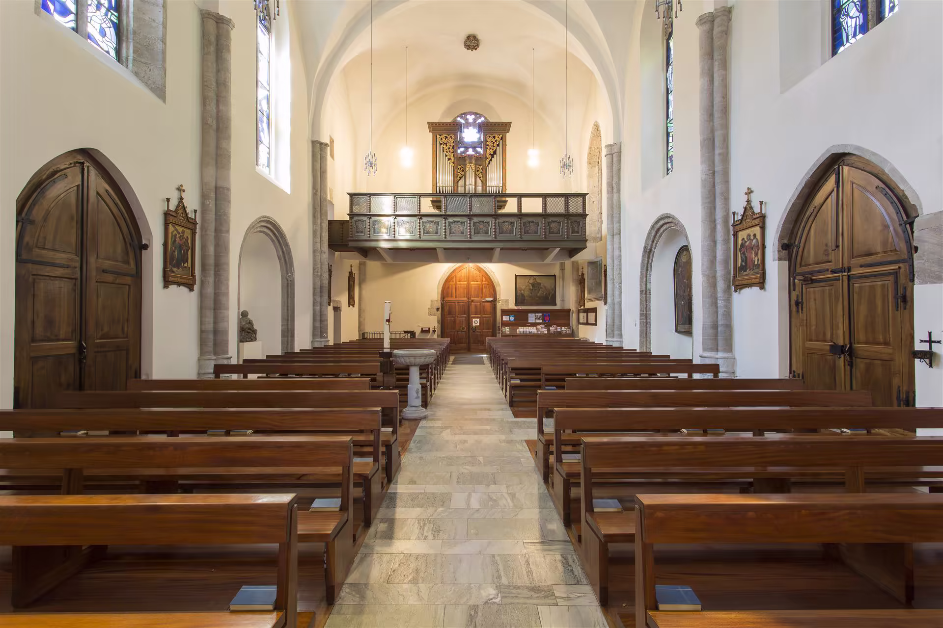 Intérieur d'une église avec des bancs en bois alignés, un orgue en hauteur, portes en bois et vitraux colorés aux murs blancs.